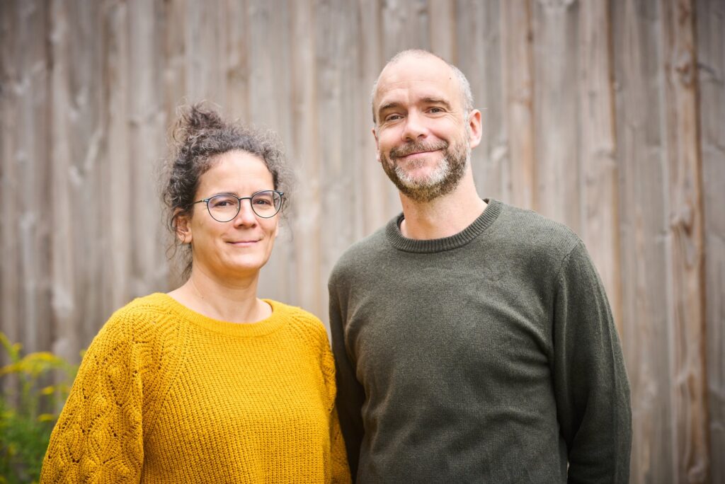 Barbara Roß und Sven Trogus vor einer Holzwand, lachend